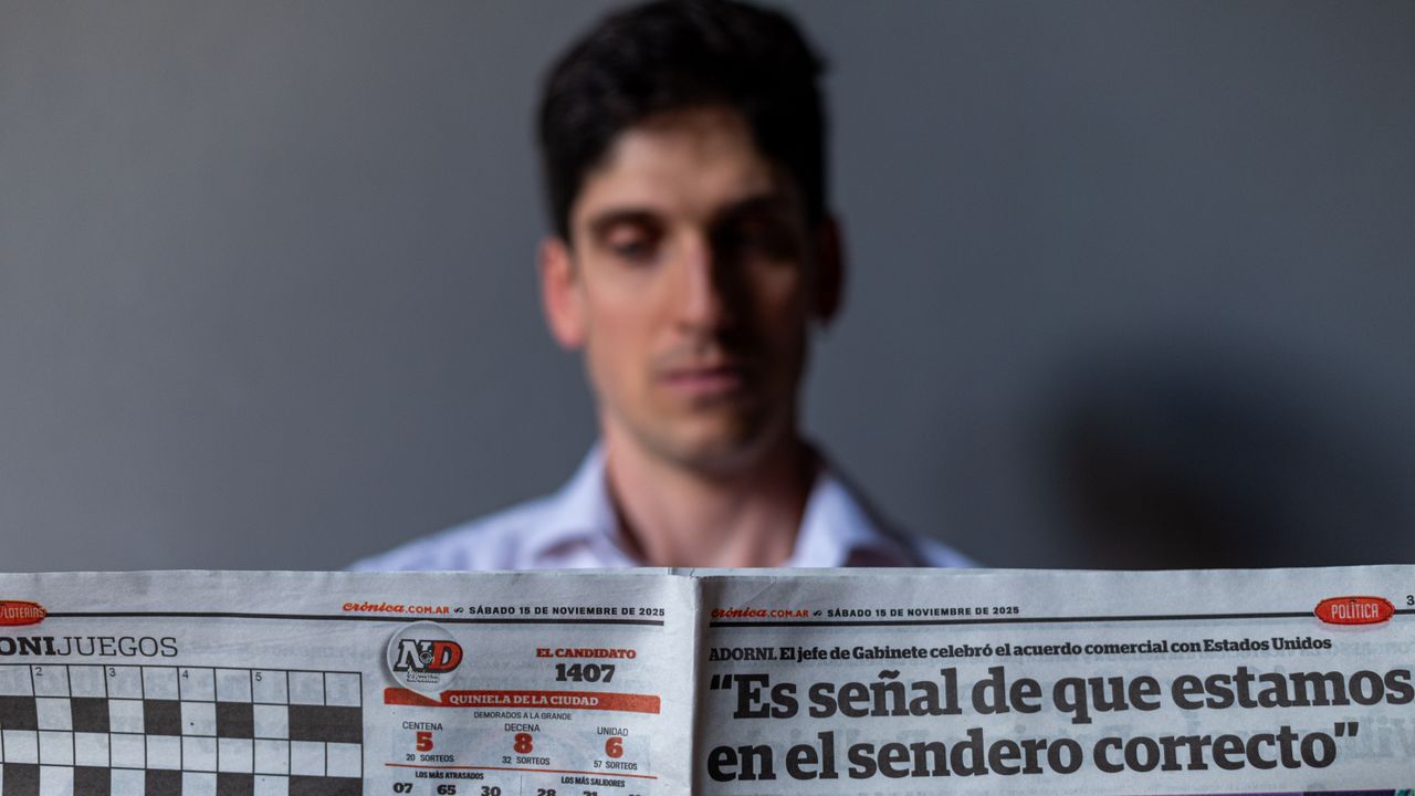 Man holding a Spanish newspaper with headline and date visible, indoors.